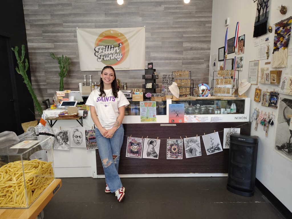 A smiling woman standing in a shop called 'Fillin' Funky', surrounded by a variety of handcrafted jewelry displayed on a counter. The shop features artwork on the walls, including sketches and photographs, with a bright and welcoming atmosphere.