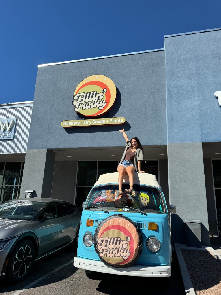 A woman sitting on top of a vintage blue Volkswagen van in front of a store sign that reads 'Fillin' Funky' featuring the tagline 'Refillery + Dry Goods + Plants'. Clear blue sky in the background.