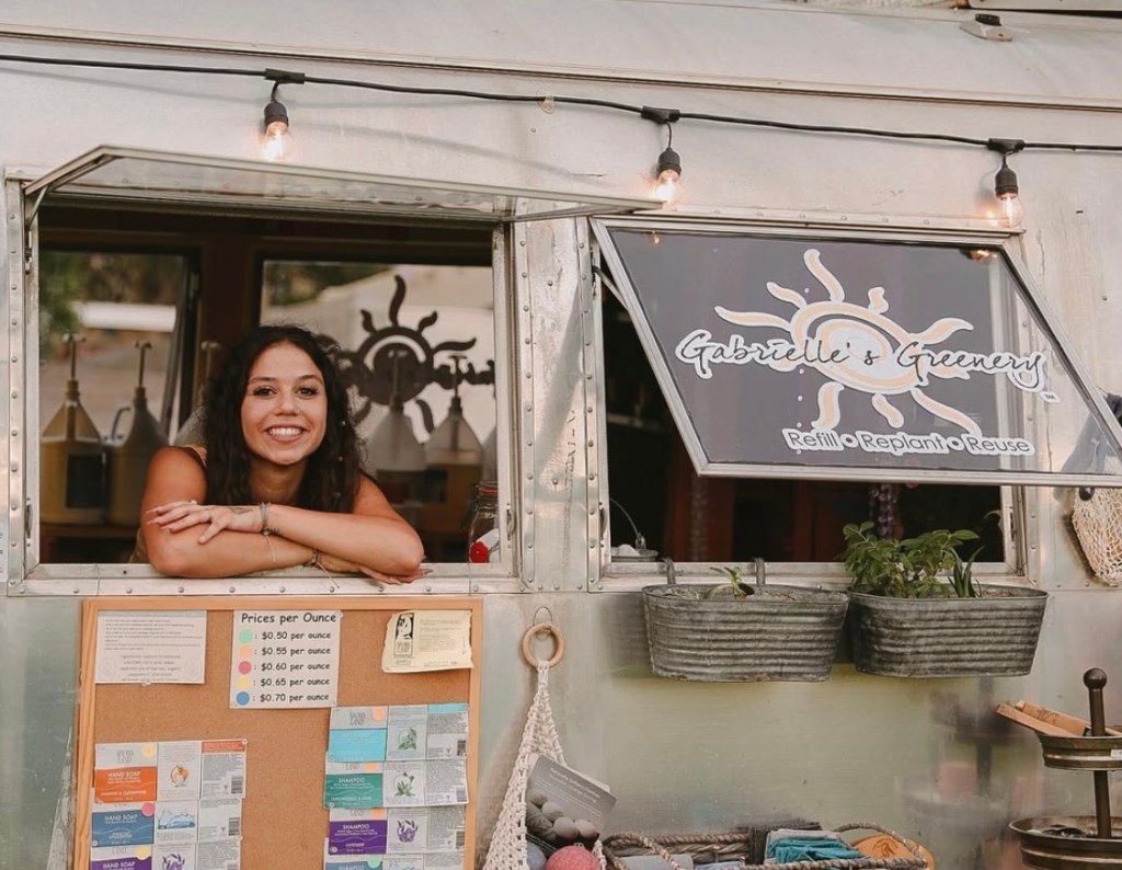 A smiling woman with curly hair leans out of a window of a food trailer called 'Gabrielle's Greenery', which promotes sustainable practices. The window displays a sign with the slogan 'Refill • Replant • Reuse'.