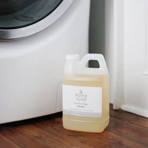 A container of Mama Suds laundry soap positioned next to a washing machine on wooden flooring.