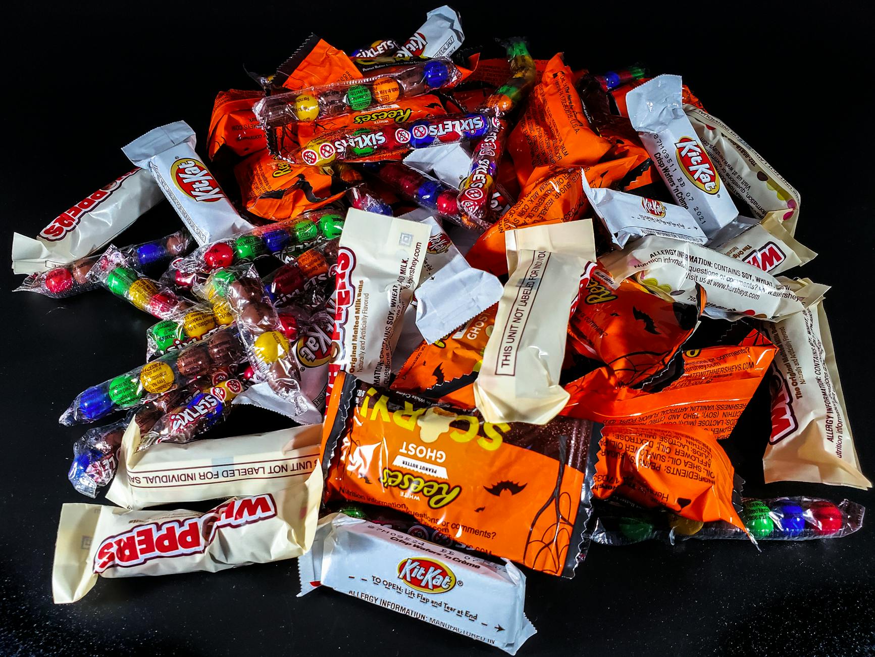 A colorful assortment of Halloween candies, including KitKats, Reese's, Butterfingers, and Skittles, arranged in a pile on a dark surface.