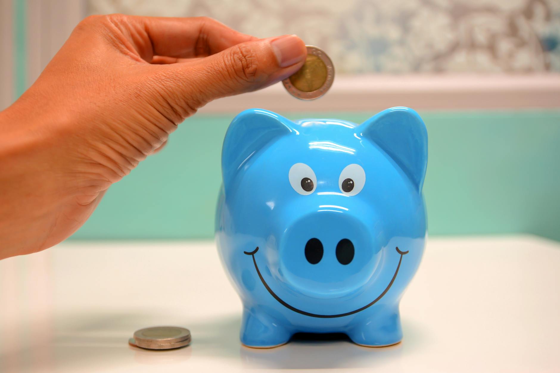 A person places a coin into a blue piggy bank with a smiley face, while another coin is visible beside it.