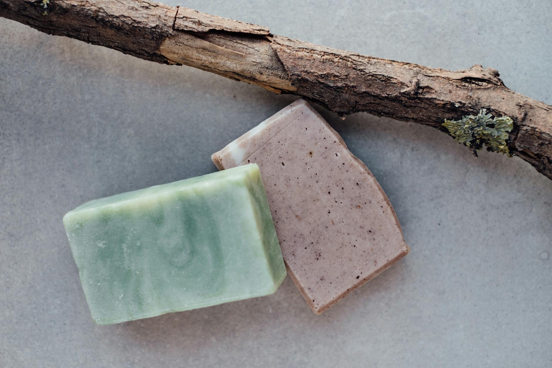 Two bars of handmade soap, one green and swirled, the other a light brown with speckles, placed next to a piece of natural wood.