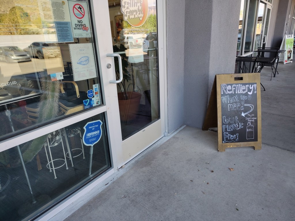 Exterior view of a storefront with a sign promoting a refillery that emphasizes zero waste and being plastic free. The door displays multiple signs, including a security sticker and a 'closed' notice.